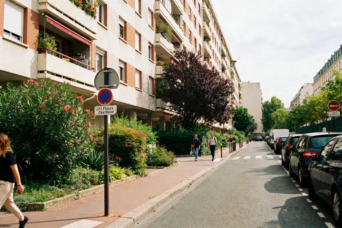 Rue résidentielle bordée d'arbres, immeuble, voitures stationnées, deux personnes marchent sur le trottoir à Courbevoie.