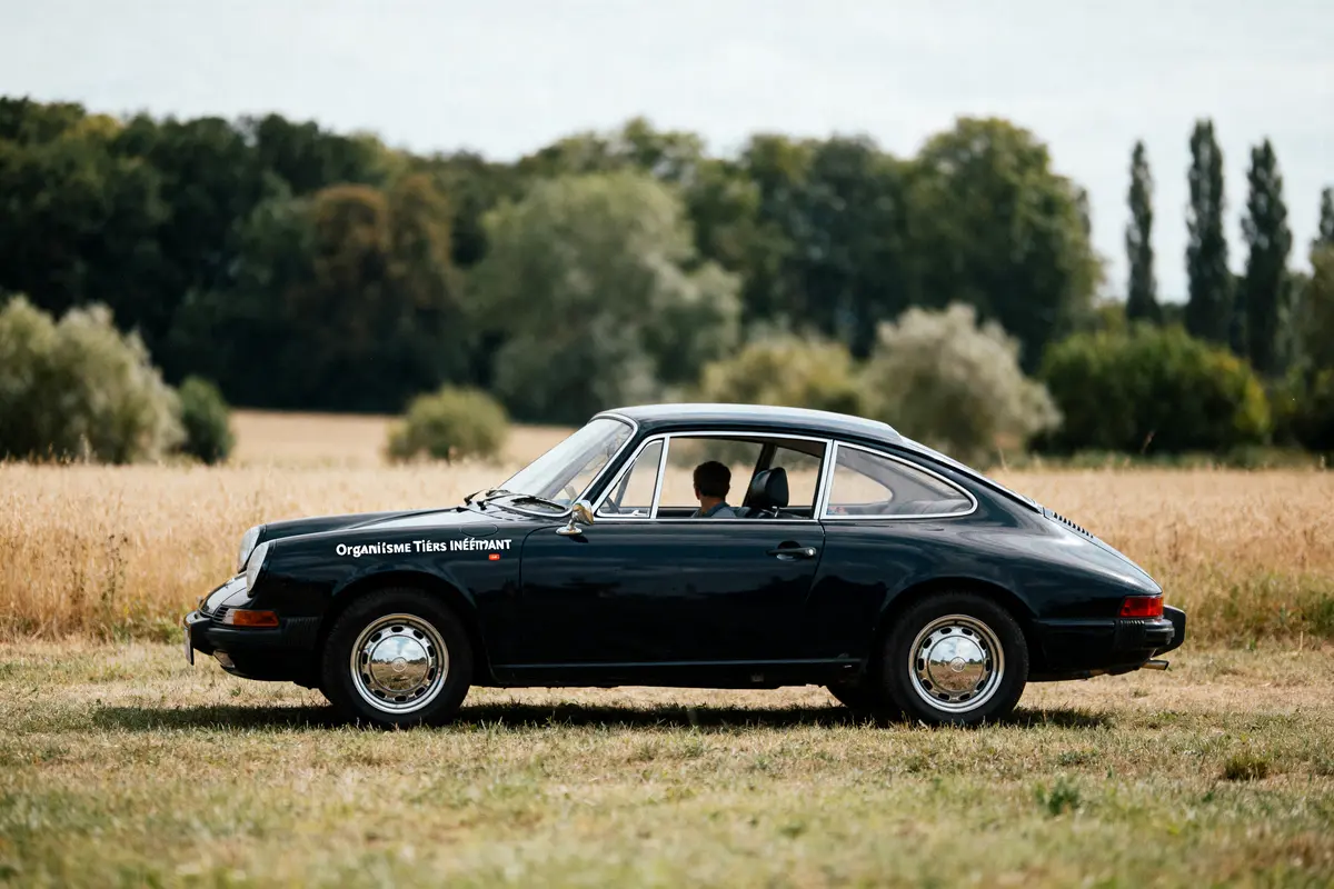 Voiture classique noire garée dans un champ, arbres en arrière-plan, homme assis à l’intérieur.