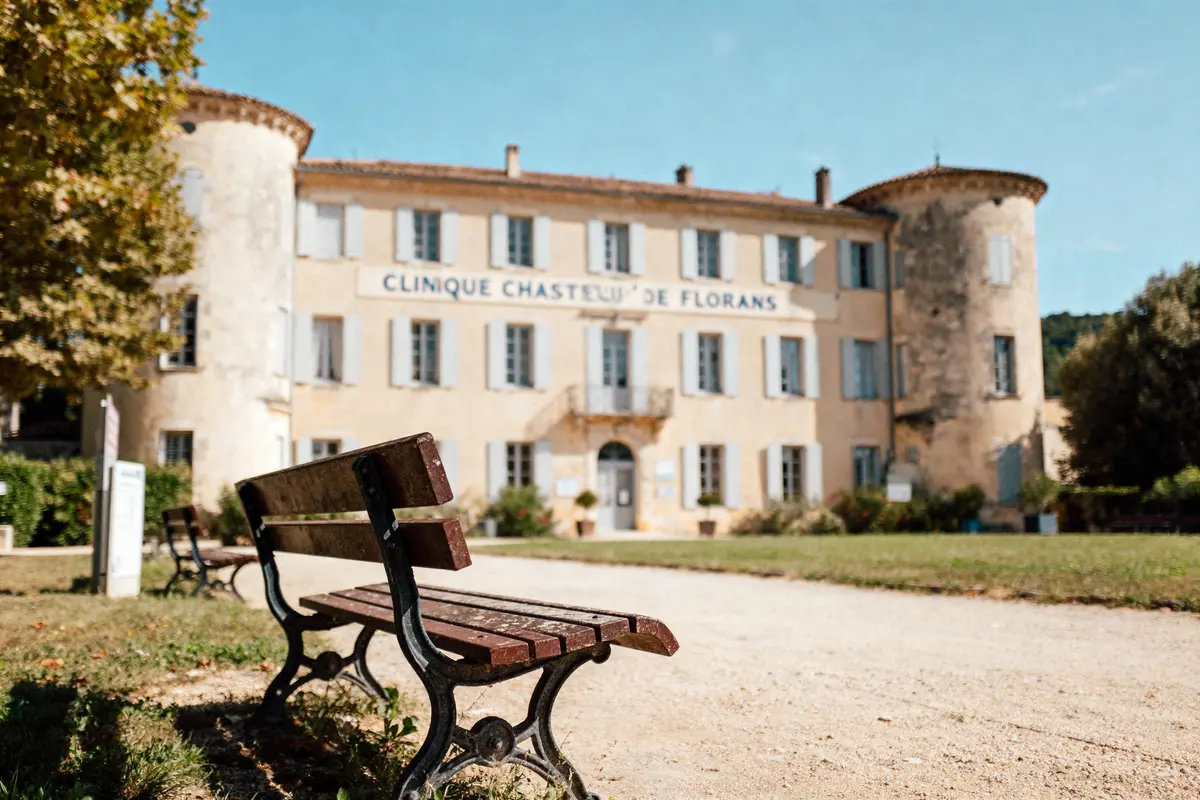 Banc dans un parc devant Clinique Château de Florans, bâtiment historique avec tours rondes, ciel bleu.