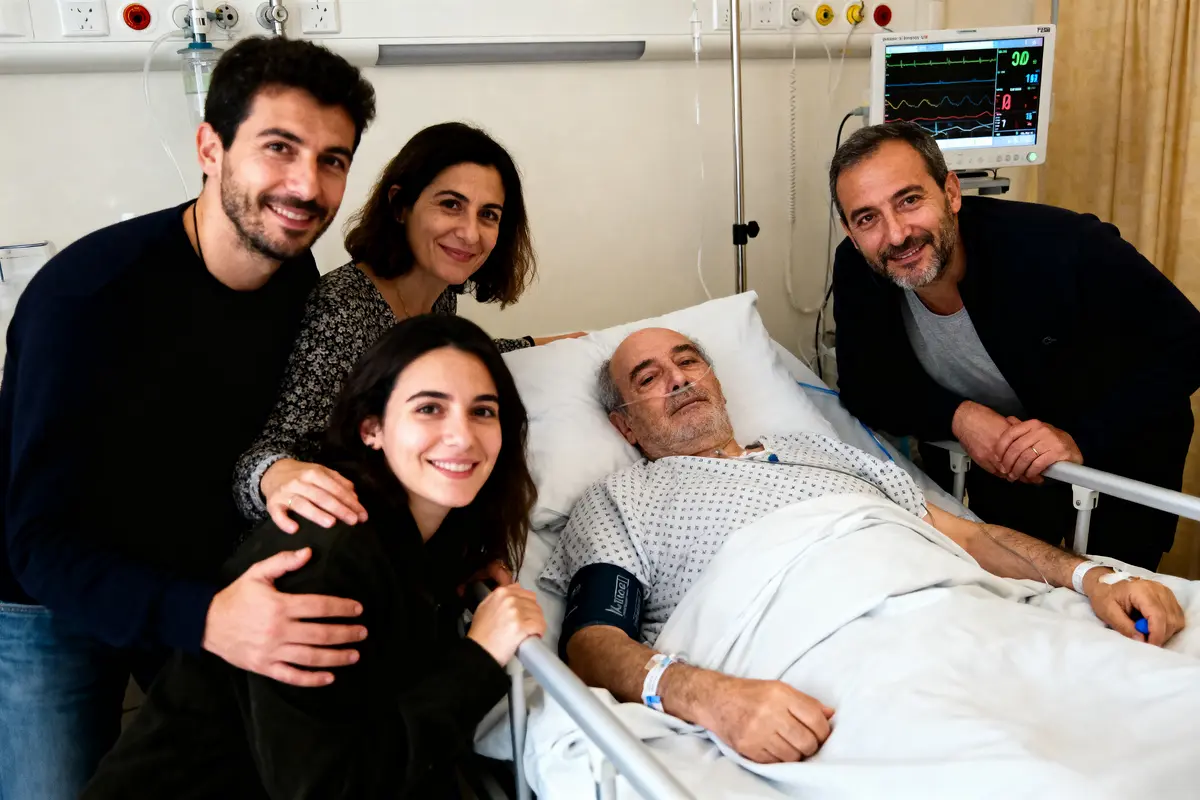 Un homme âgé alité entouré de quatre personnes souriantes à l'hôpital, avec moniteur de santé visible.