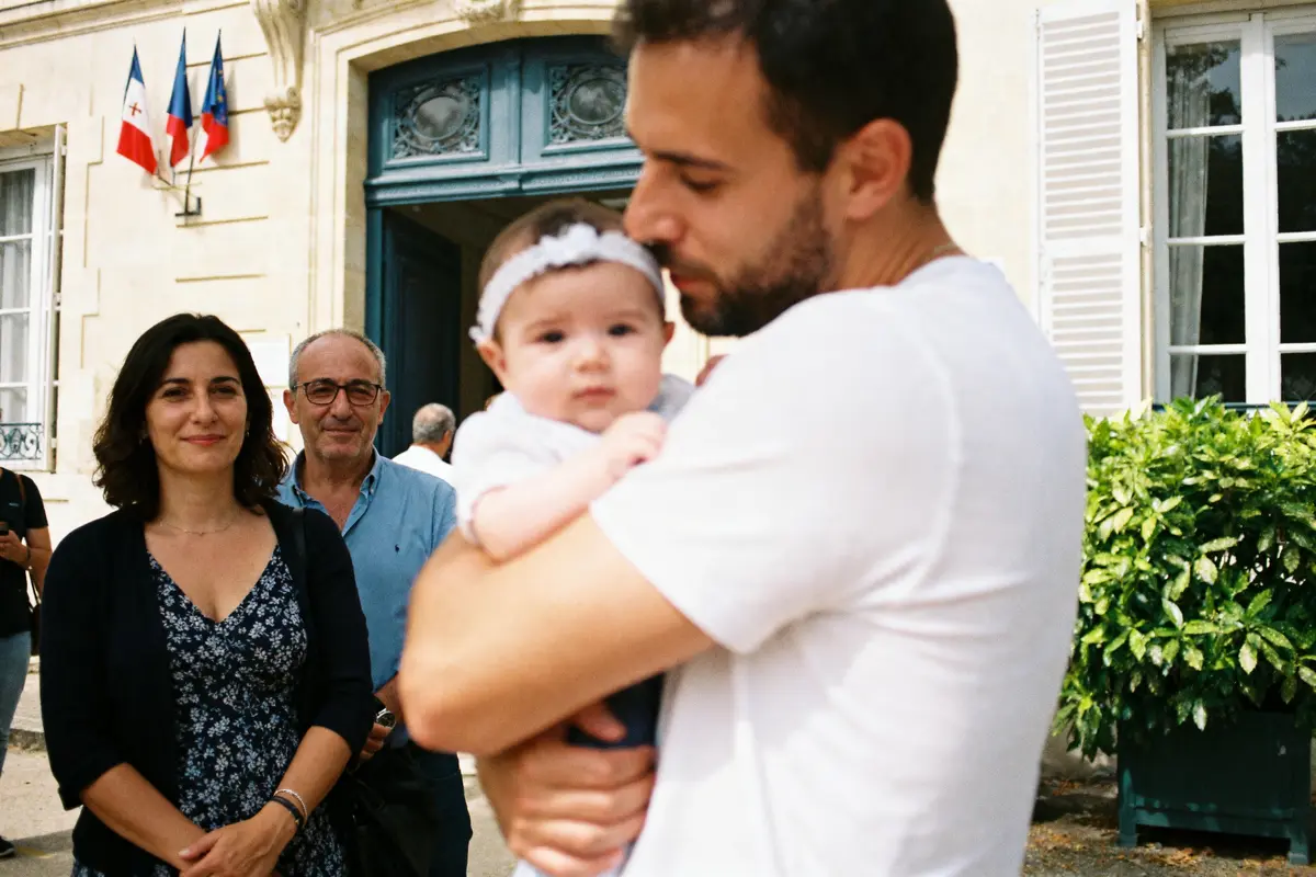 Homme tenant un bébé, avec deux personnes en arrière-plan devant un bâtiment, drapeaux français visibles.